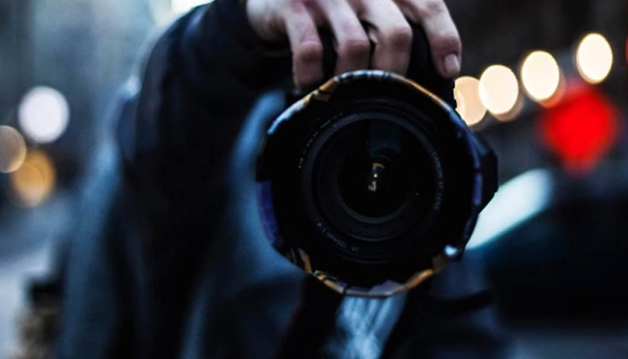 Fotografía en primer plano del objetivo de la cámara y mano del fotógrafo realizada frente a un espejo. El punto de enfoque permite ver ese reflejo mientras el fondo de la ciudad queda totalmente desenfocado. Se pueden apreciar reflejos de luces y poco más.