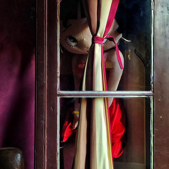 Fotografía del pelucha a tamaño real del personaje Betty Boop escondida detrás de las puertas de una ventana de madera con cristales. Un juego visual donde Betty parece decir uuups...