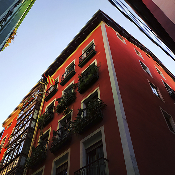 Fotografía contrapicada de unos edificios pintados con colores cálidos. Los vértices de las fachadas parecen proyectarse hacia el cielo. En uno de esos balcones se aprecian varias banderas. En territorio Vasco comparte su bandera espacio con la bandera catalana independentista.